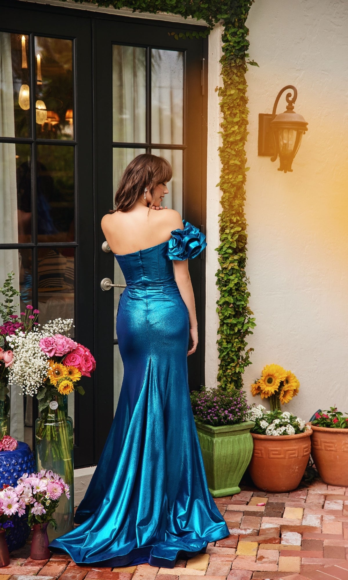 A woman in the Ava Presley 40082 Long Ruffle Metallic Prom Dress stands on a brick patio facing away, surrounded by potted flowers and greenery outside a glass door.